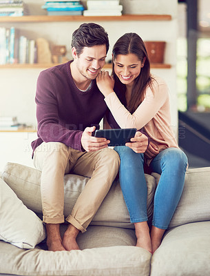 Buy stock photo Shot of a cheerful young couple using a digital tablet while relaxing on the sofa at home