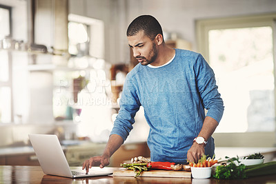 Buy stock photo Young man using a laptop while cooking in a kitchen, checking the internet. Male watching a tutorial online while preparing a healthy, organic homemade meal. Guy following a easy vegetarian recipe