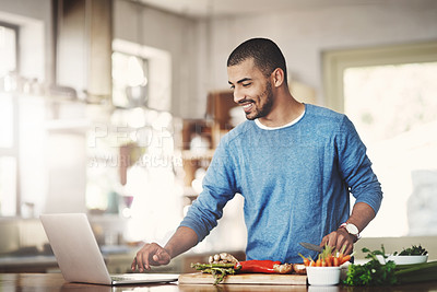 Buy stock photo Young casual man in the kitchen on a laptop while preparing a healthy meal at home. Happy smiling male browsing and learning on computer on how to cook. Guy alone checking online recipes on the web.