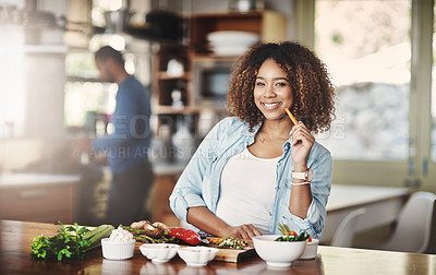 Buy stock photo Portrait of a young woman holding a carrot while preparing a healthy meal with her husband in the background