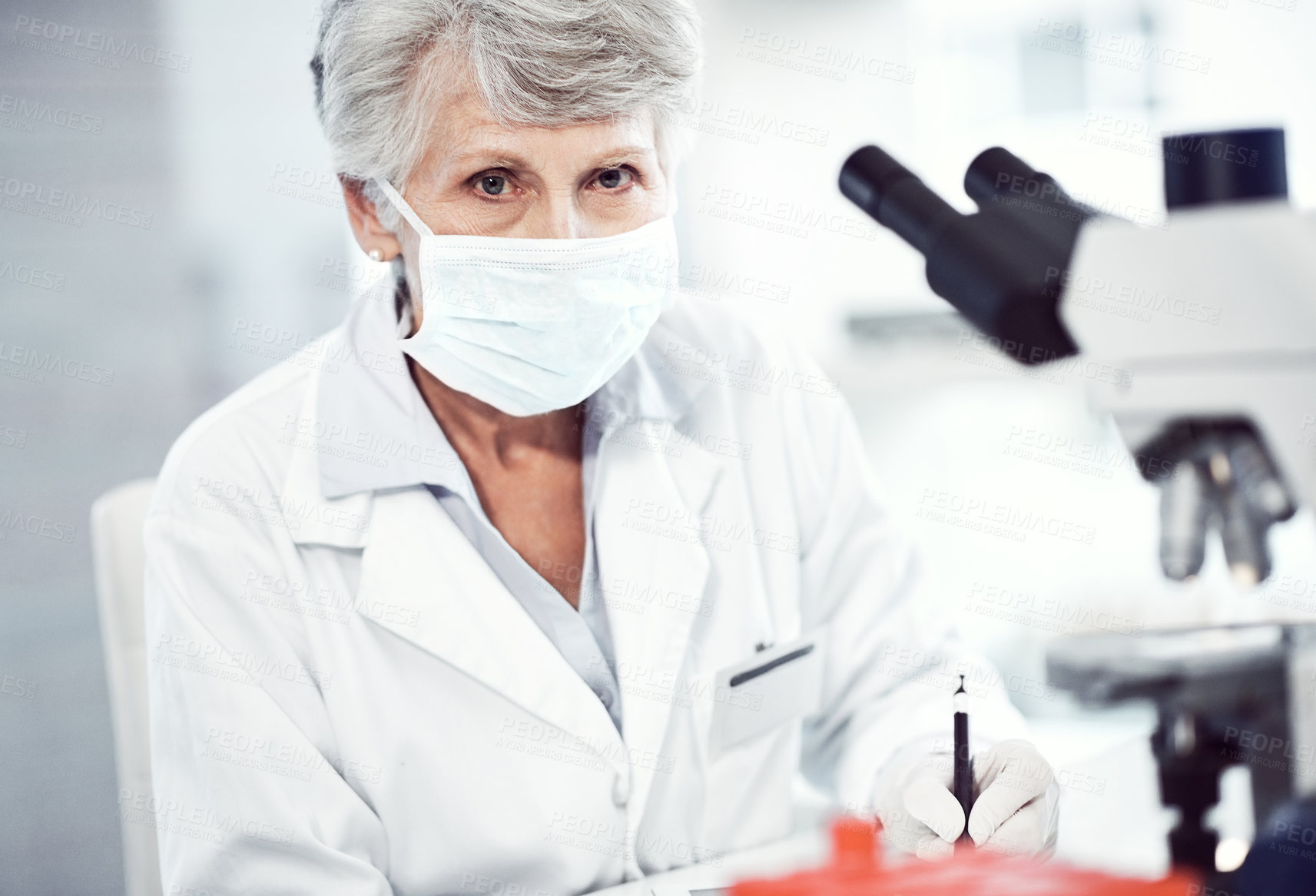 Buy stock photo Portrait of a focused elderly female scientist making notes while looking into the camera and wearing a doctor's mask inside of a laboratory