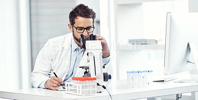 Buy stock photo Shot of a focused young male scientist making notes while looking trough a microscope and being seated inside a laboratory