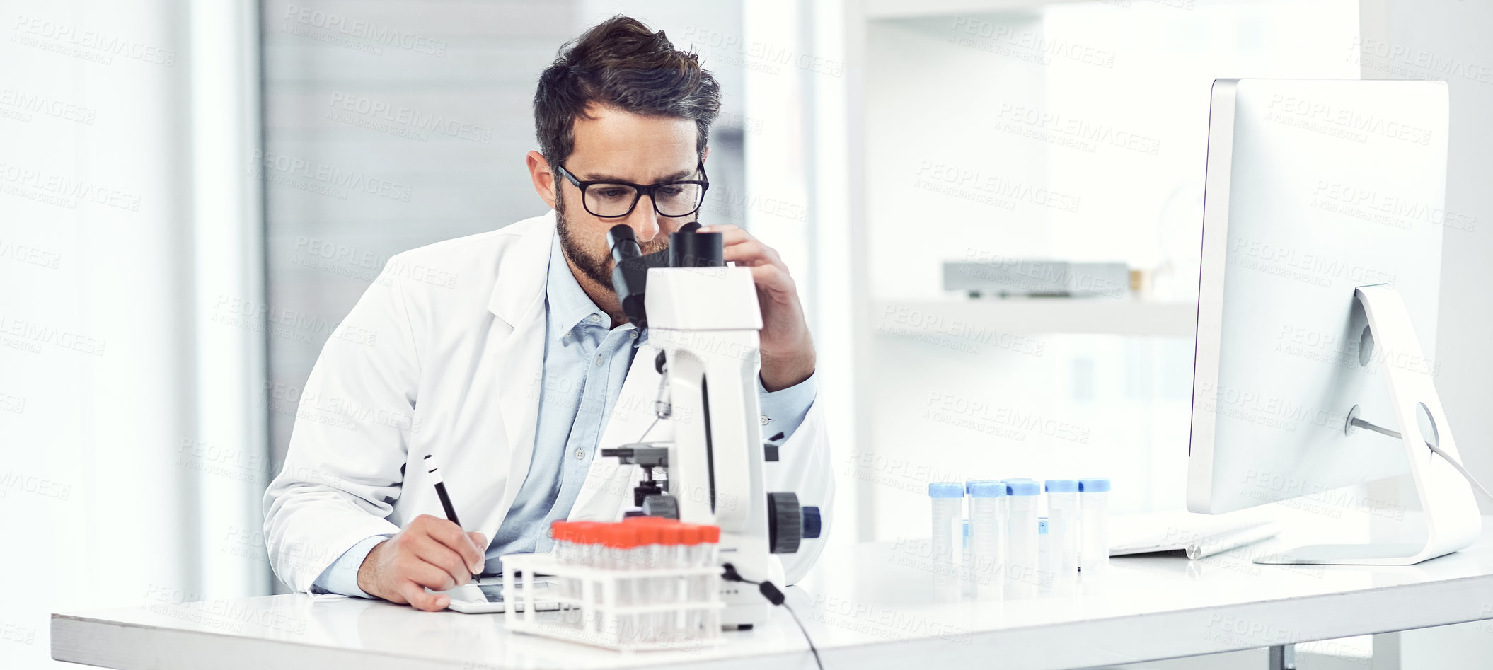 Buy stock photo Shot of a focused young male scientist making notes while looking trough a microscope and being seated inside a laboratory