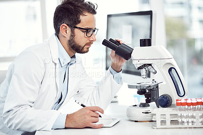 Buy stock photo Shot of a focused young male scientist making notes and looking through a microscope while being seated inside of a laboratory