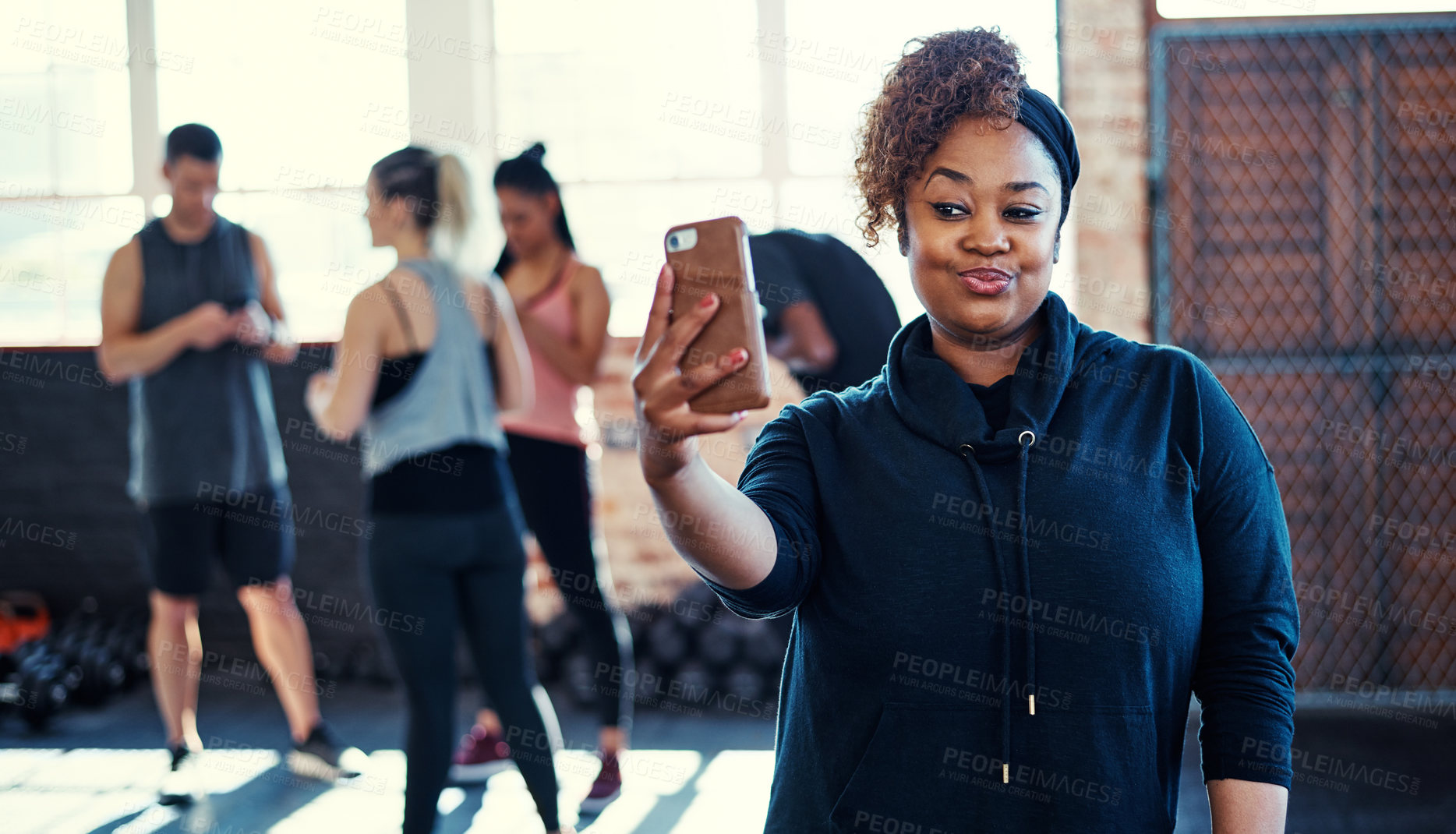 Buy stock photo Shot of a cheerful young woman standing and taking a self portrait with her cellphone before a workout in a gym