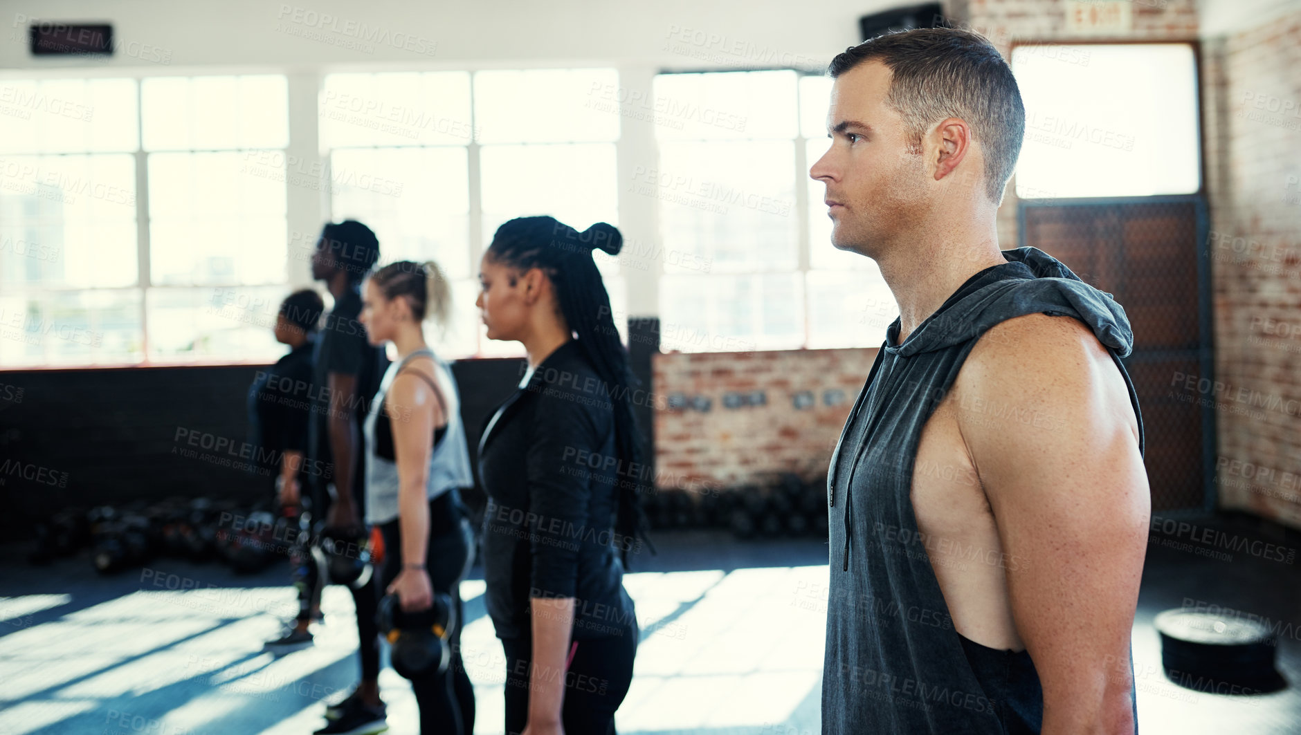 Buy stock photo Shot of a focused group of young people standing in a row and training with weights in a gym