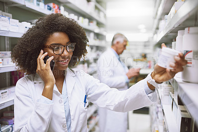 Buy stock photo Shot of a cheerful young female pharmacist talking on her cellphone while reading the back of a medication box