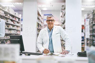 Buy stock photo Portrait of a cheerful mature male pharmacist making notes in a book on the counter while looking at the camera