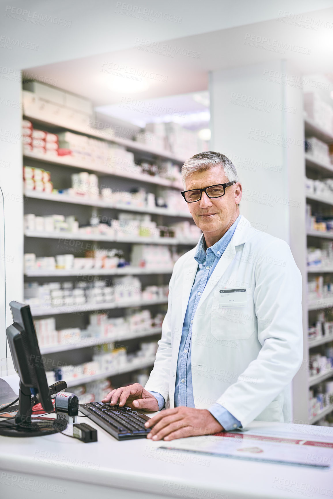 Buy stock photo Portrait of a confident mature male pharmacist typing on a computer while looking at the camera in the pharmacy