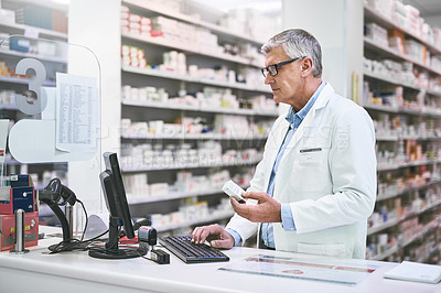 Buy stock photo Shot of a focused mature male pharmacist typing on a computer keyboard while holding a medication box