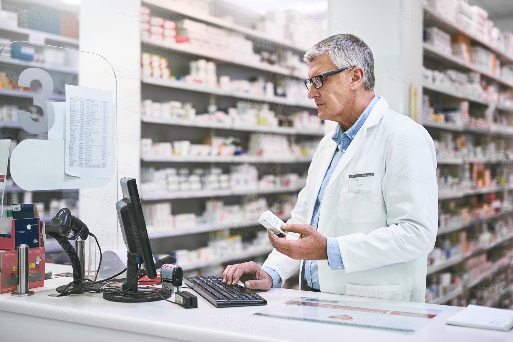 Buy stock photo Shot of a focused mature male pharmacist typing on a computer keyboard while holding a medication box