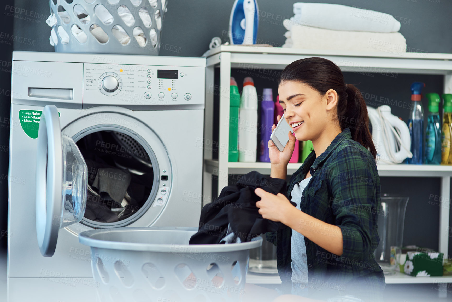 Buy stock photo Cropped shot of an attractive young woman using her cellphone while doing laundry at home