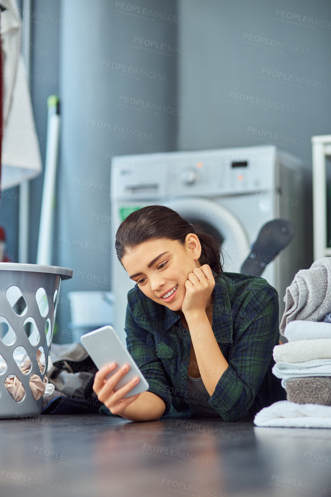 Buy stock photo Shot of an attractive young woman using her phone while lying on the floor waiting for the next load of laundry to be done