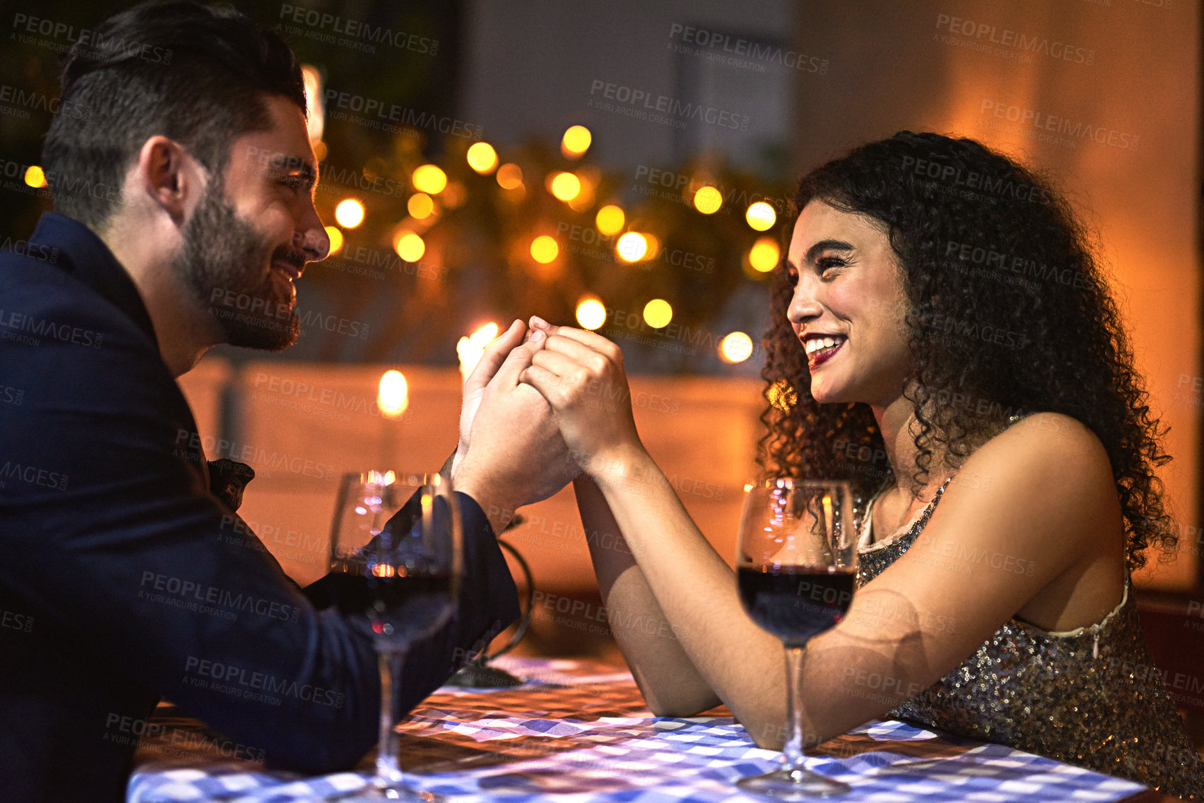Buy stock photo Shot of a cheerful young couple holding hands while looking into each other's eyes over a candle lit dinner date at night