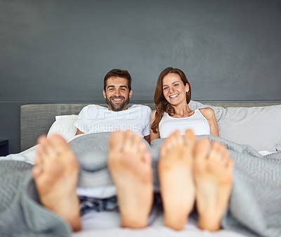 Buy stock photo Shot of a happy young couple relaxing in bed with their feet poking out from under the bed sheets