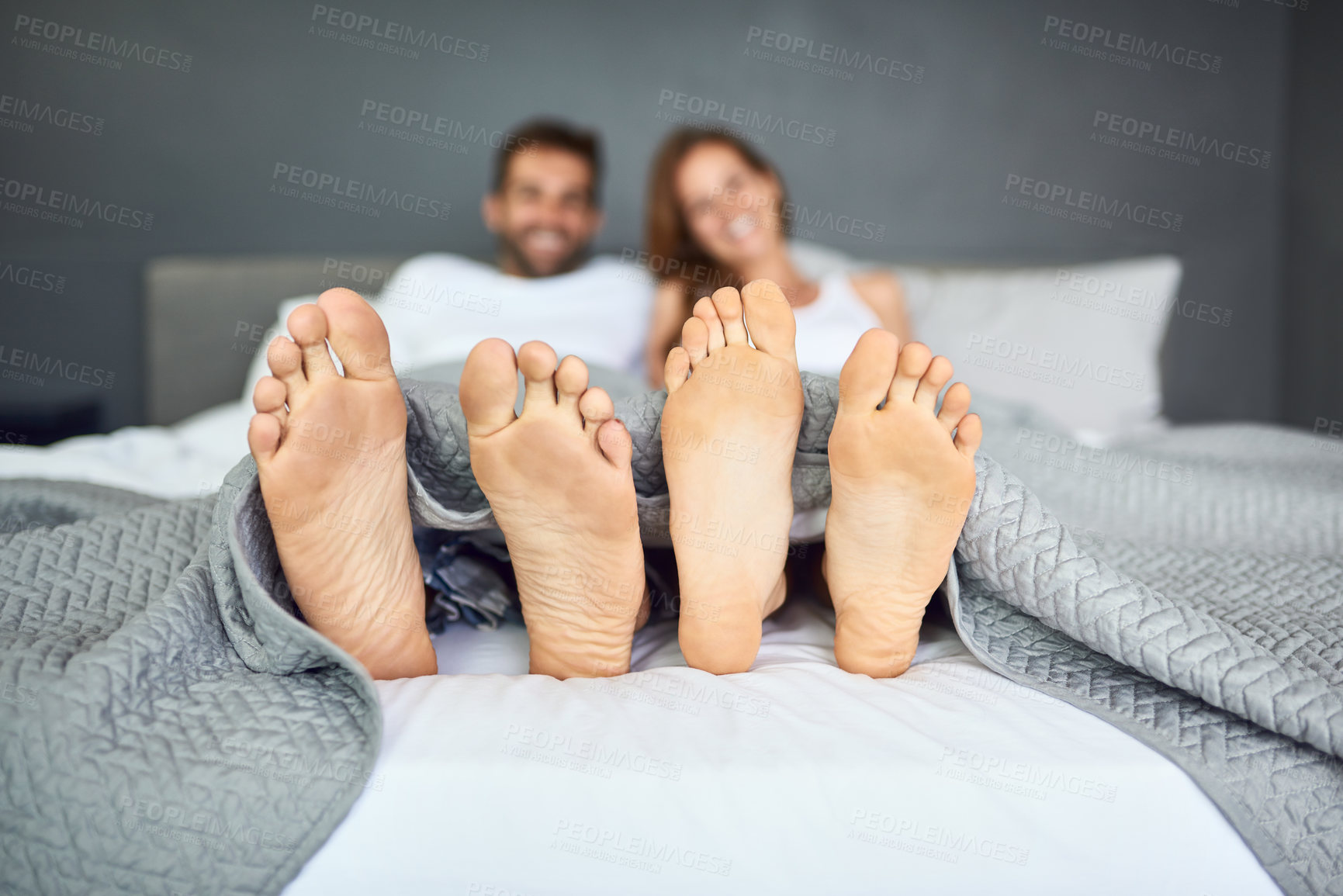 Buy stock photo Shot of a happy young couple relaxing in bed with their feet poking out from under the bed sheets