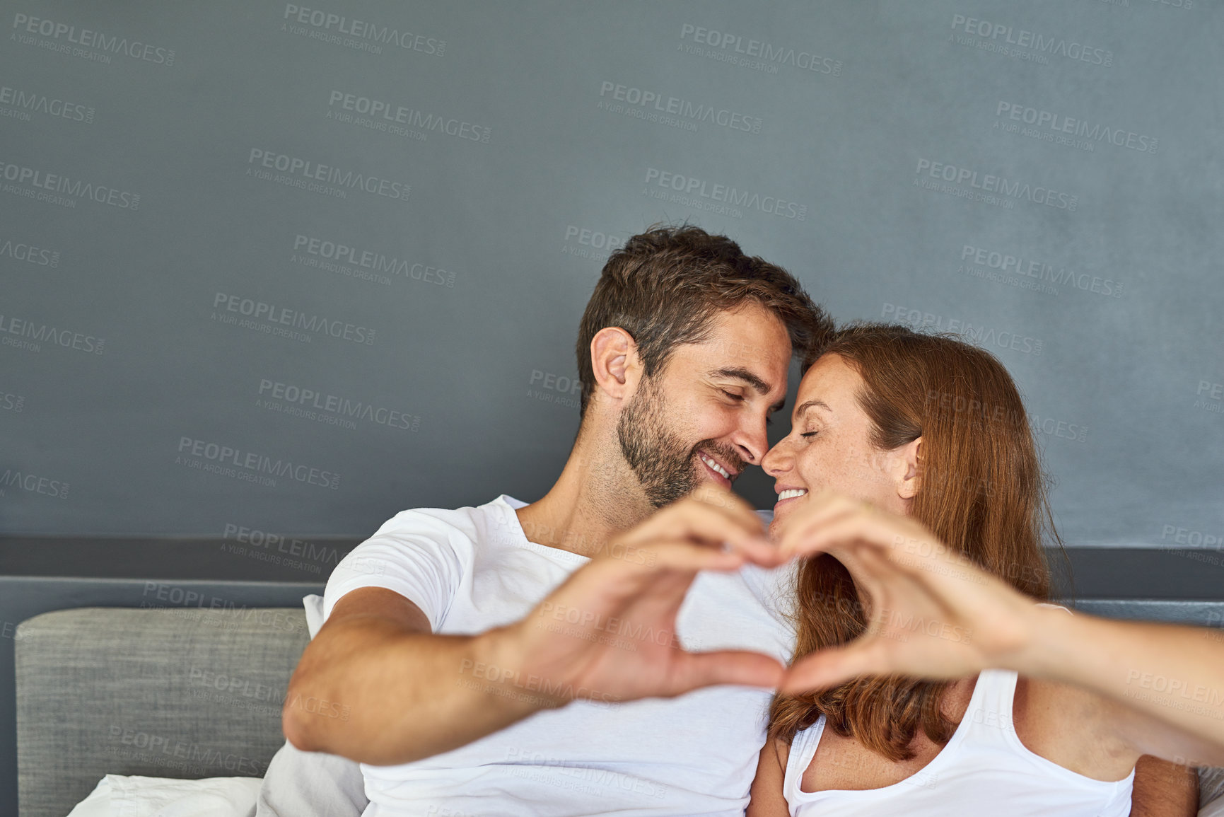Buy stock photo Shot of a happy young couple relaxing in bed and making a heart gesture with their hands