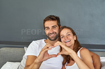 Buy stock photo Shot of a happy young couple relaxing in bed and making a heart gesture with their hands