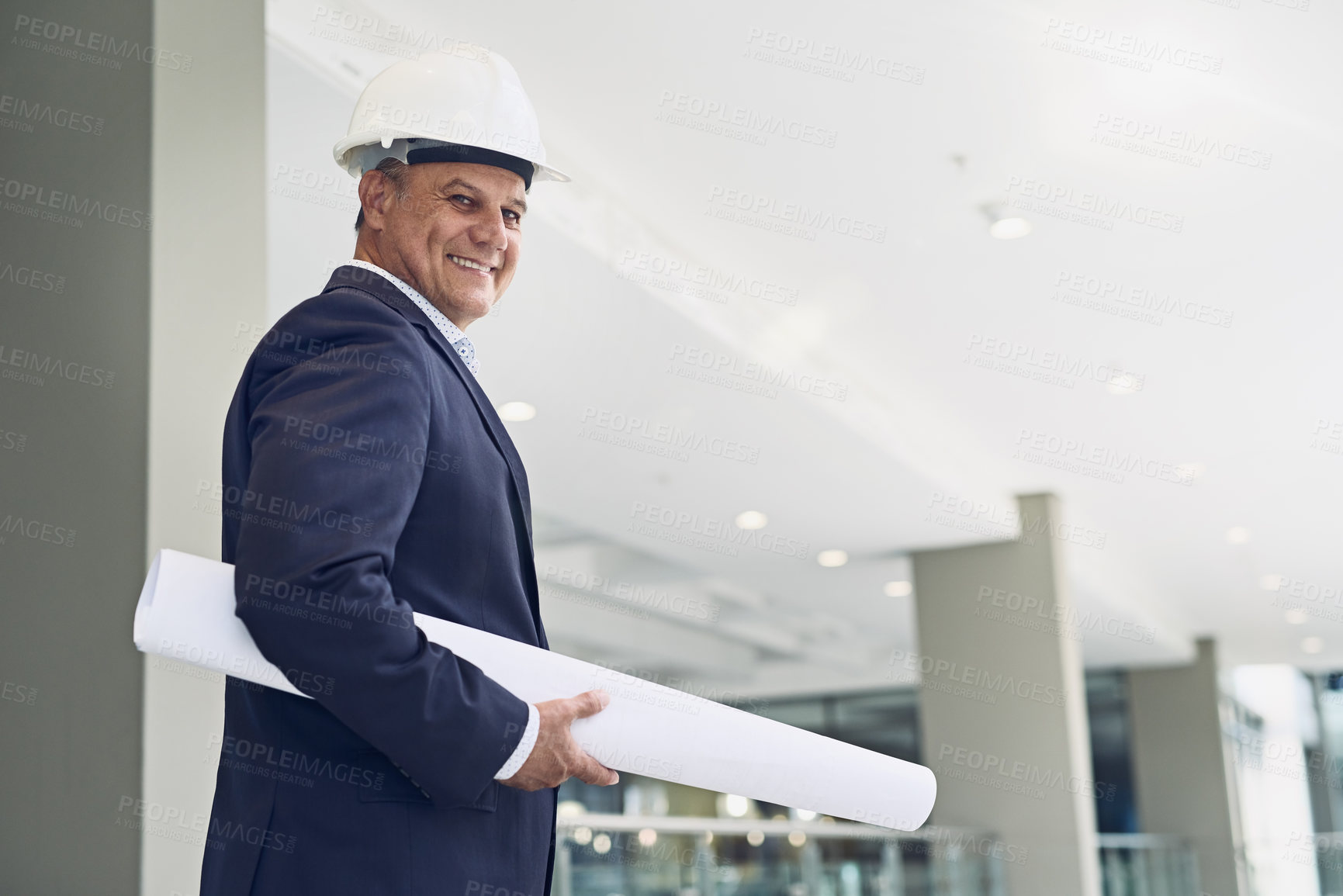 Buy stock photo Portrait of a cheerful professional male architect looking at the camera while holding blueprints inside a building