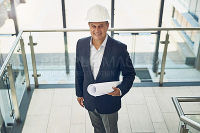 Buy stock photo Portrait of a cheerful professional male architect looking at the camera while holding blueprints inside a building