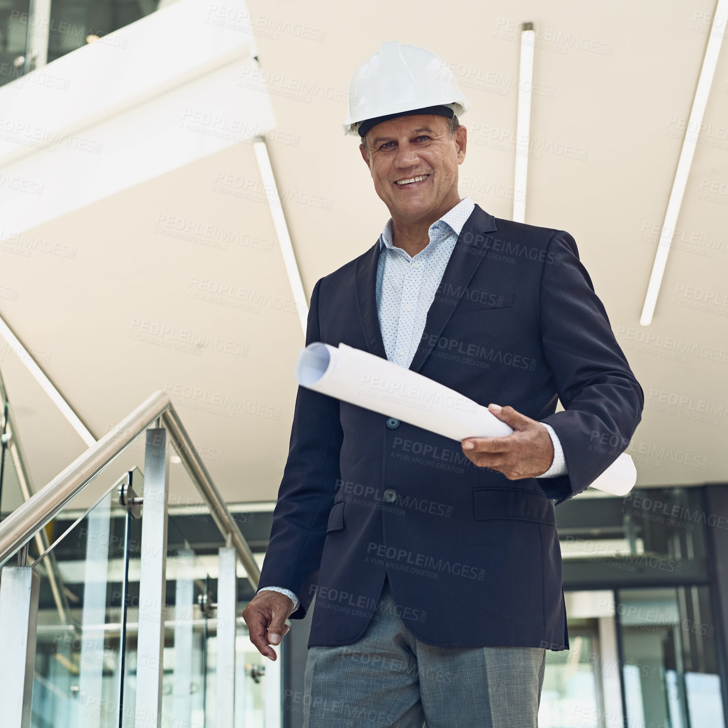 Buy stock photo Portrait of a cheerful professional male architect looking at the camera while holding blueprints inside a building