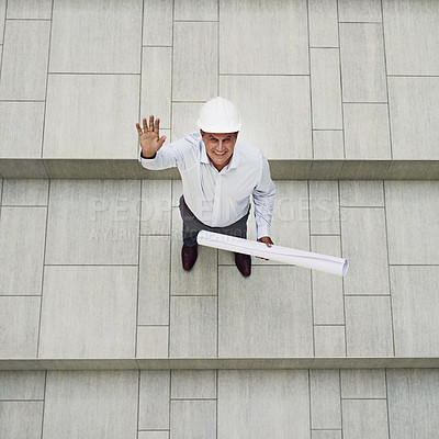 Buy stock photo High angle shot of a cheerful professional male architect holding blueprints while looking up to the camera inside of a building