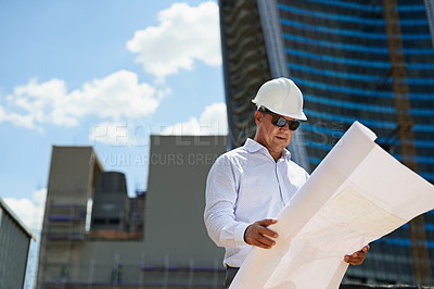 Buy stock photo Shot of a professional male architect standing on a building site while holding and reading blueprints outside