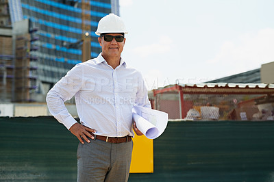 Buy stock photo Portrait of a professional male architect standing on a building site while holding blueprints and looking at the camera outside