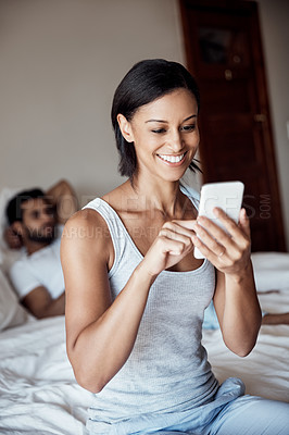 Buy stock photo Shot of a young woman using her cellphone while sitting on a bed with her husband in the background