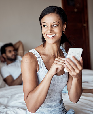 Buy stock photo Shot of a young woman using her cellphone while sitting on a bed with her husband in the background