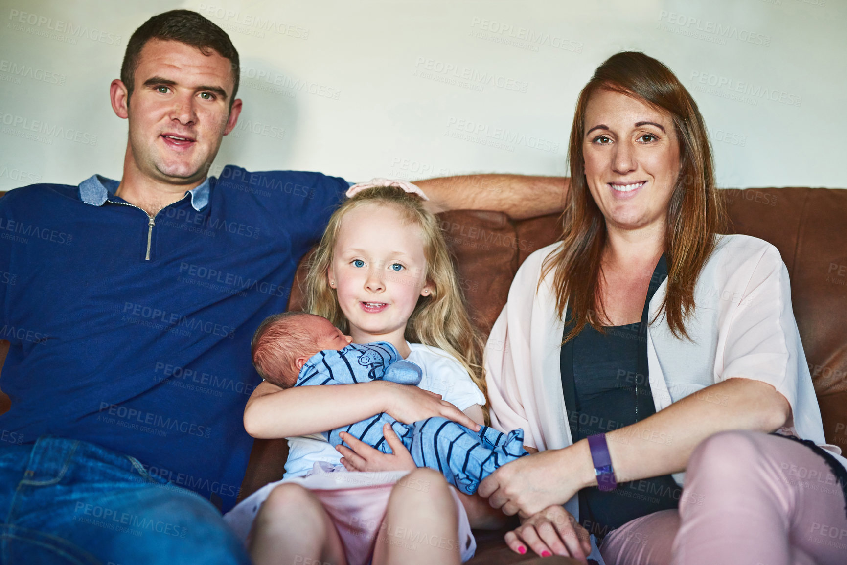 Buy stock photo Portrait of a cheerful young family grouped together on the couch while looking into the camera at home