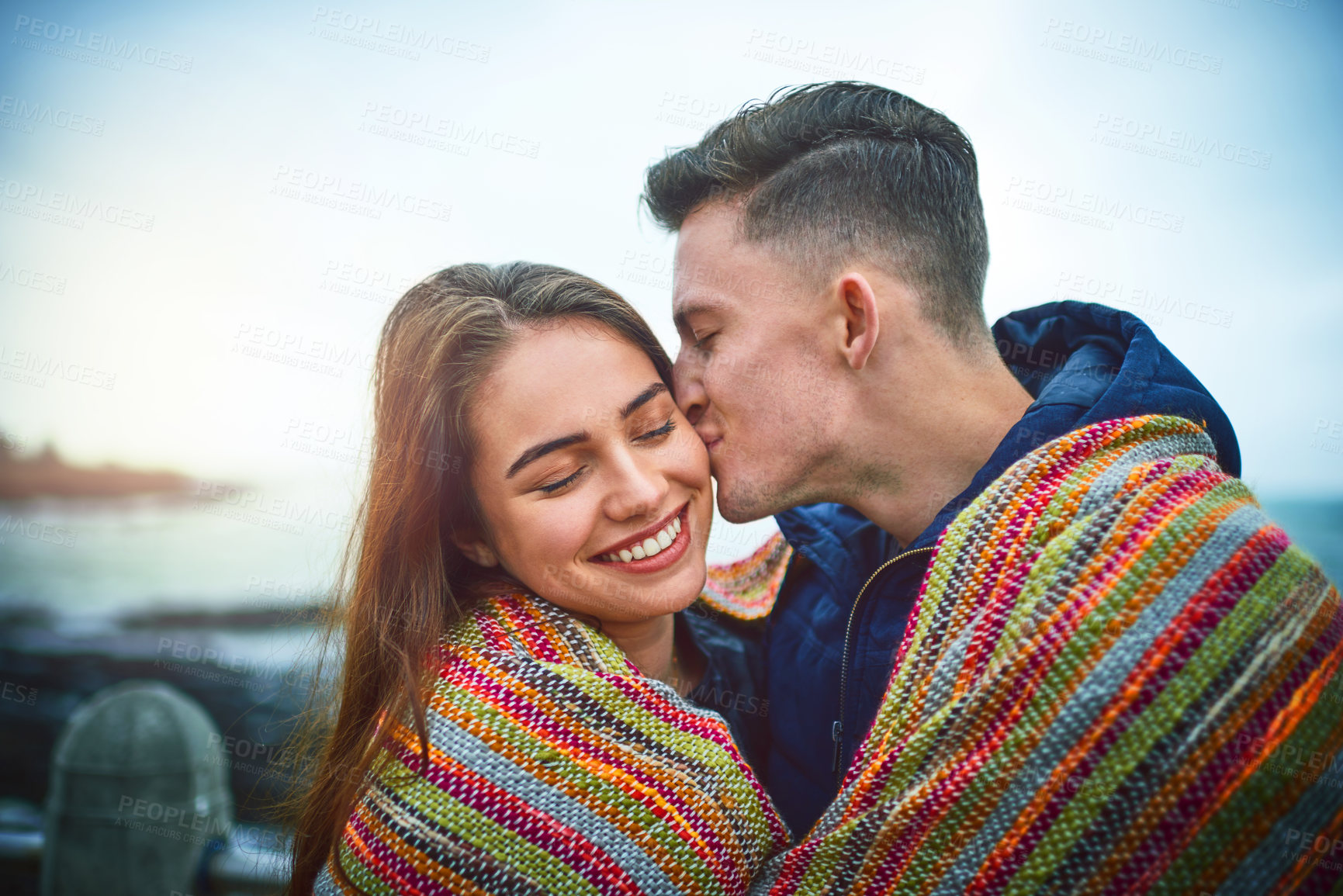 Buy stock photo Happy, couple and kiss with blanket outdoor for warmth support, summer travel and bonding together at beach. Smile, woman and man hug with romance date, winter comfort and relationship love at ocean