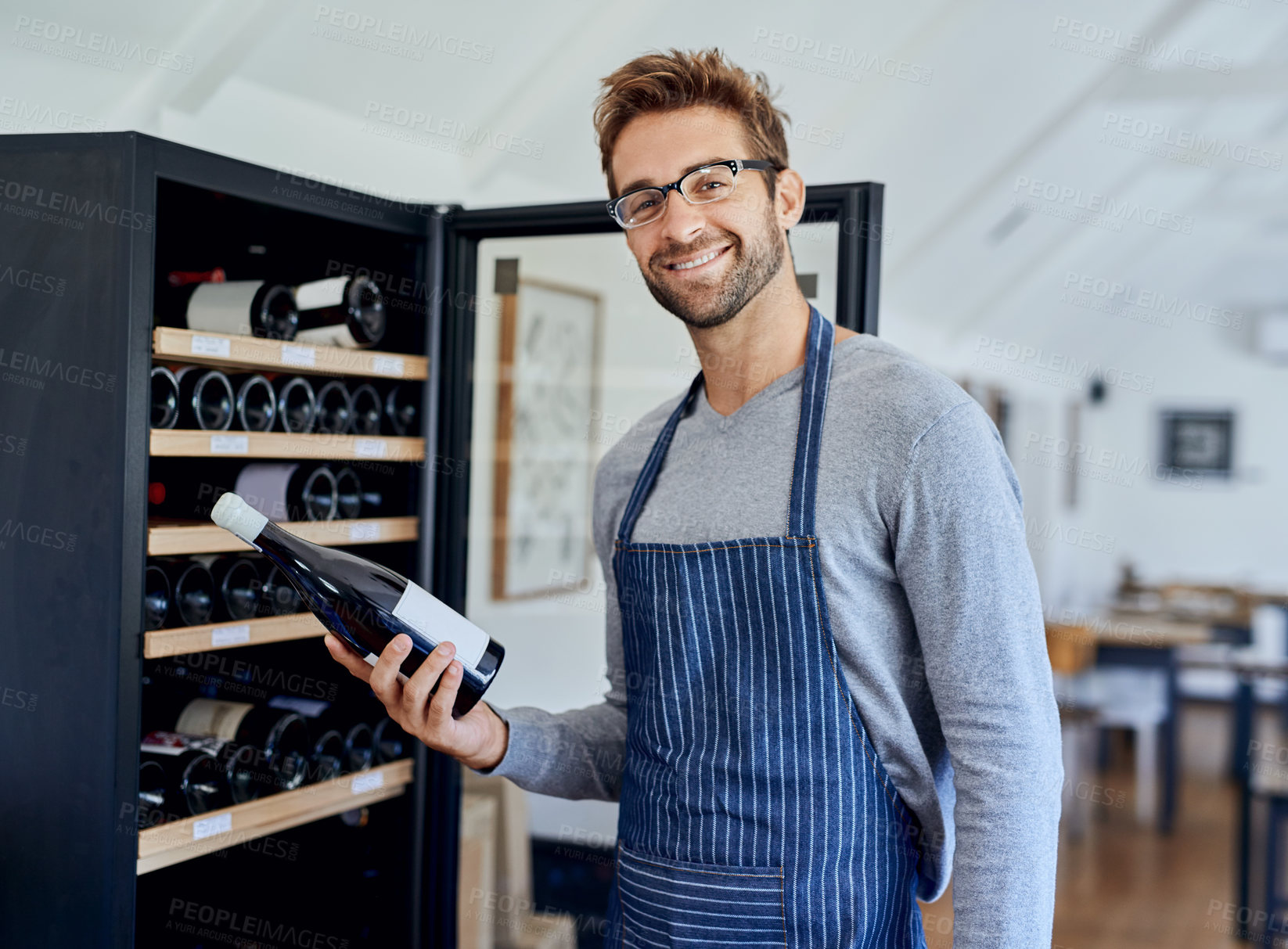 Buy stock photo Bottle, cafe and portrait of man at wine shelf for selection, choice and hospitality. Smile, alcohol and restaurant sommelier, waiter or small business owner checking drinks for service quality.