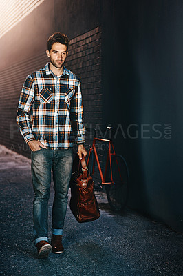 Buy stock photo Shot of a handsome young man carrying a bag and walking down a city street