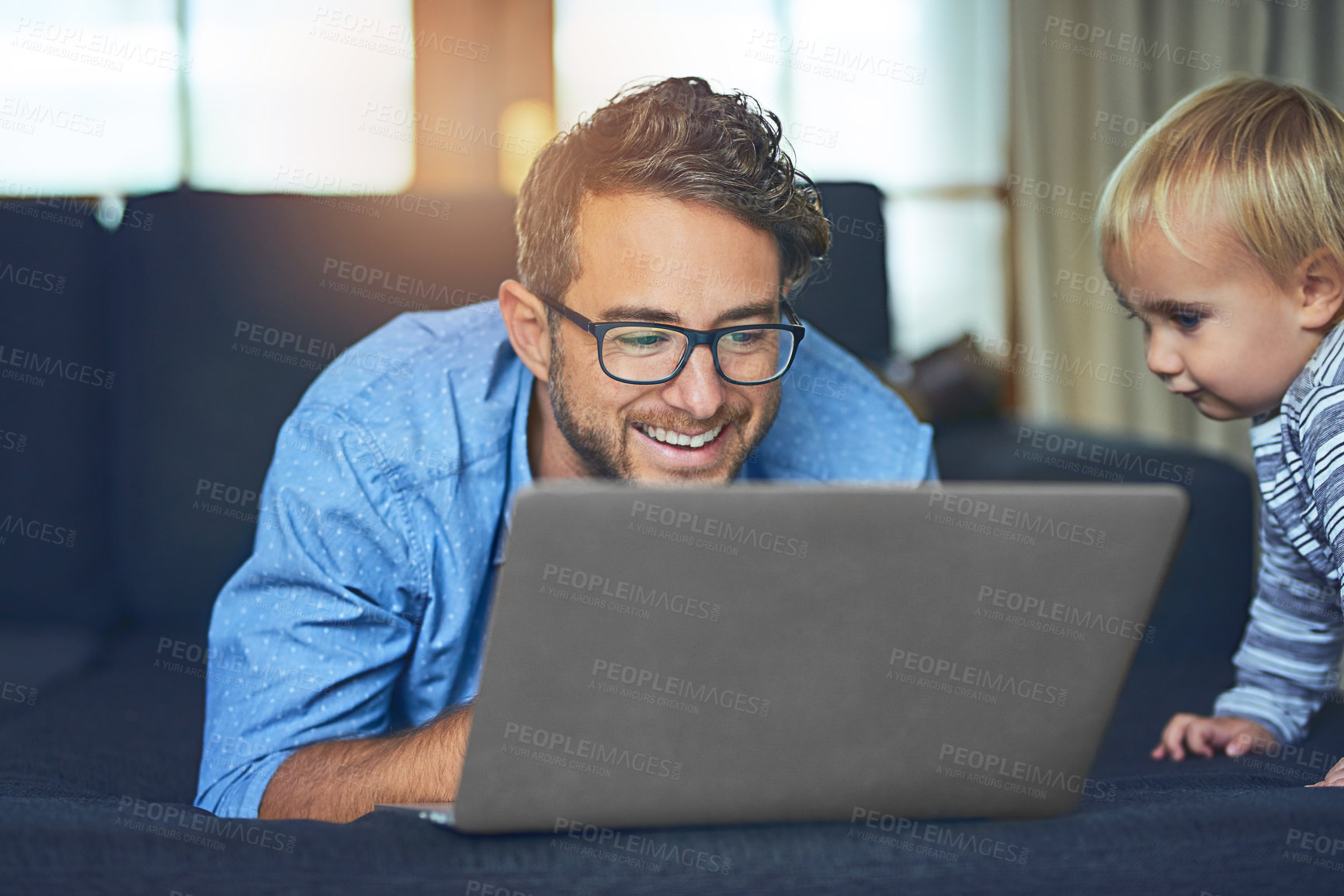 Buy stock photo Shot of a young man using a laptop on the sofa at home with his son next to him