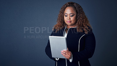 Buy stock photo Studio shot of a confident young businesswoman working on her digital tablet while standing against a blue background