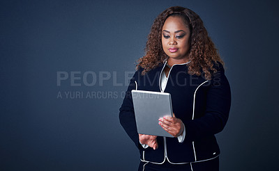 Buy stock photo Studio shot of a confident young businesswoman working on her digital tablet while standing against a blue background