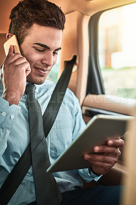Buy stock photo Shot of a young businessman talking on a phone while using a digital tablet in the back seat of a car