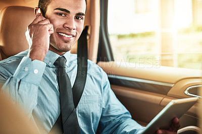 Buy stock photo Portrait of a young businessman talking on a phone while using a digital tablet in the back seat of a car