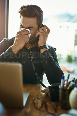 Buy stock photo Shot of a young businessman blowing his nose while speaking on a phone in an office