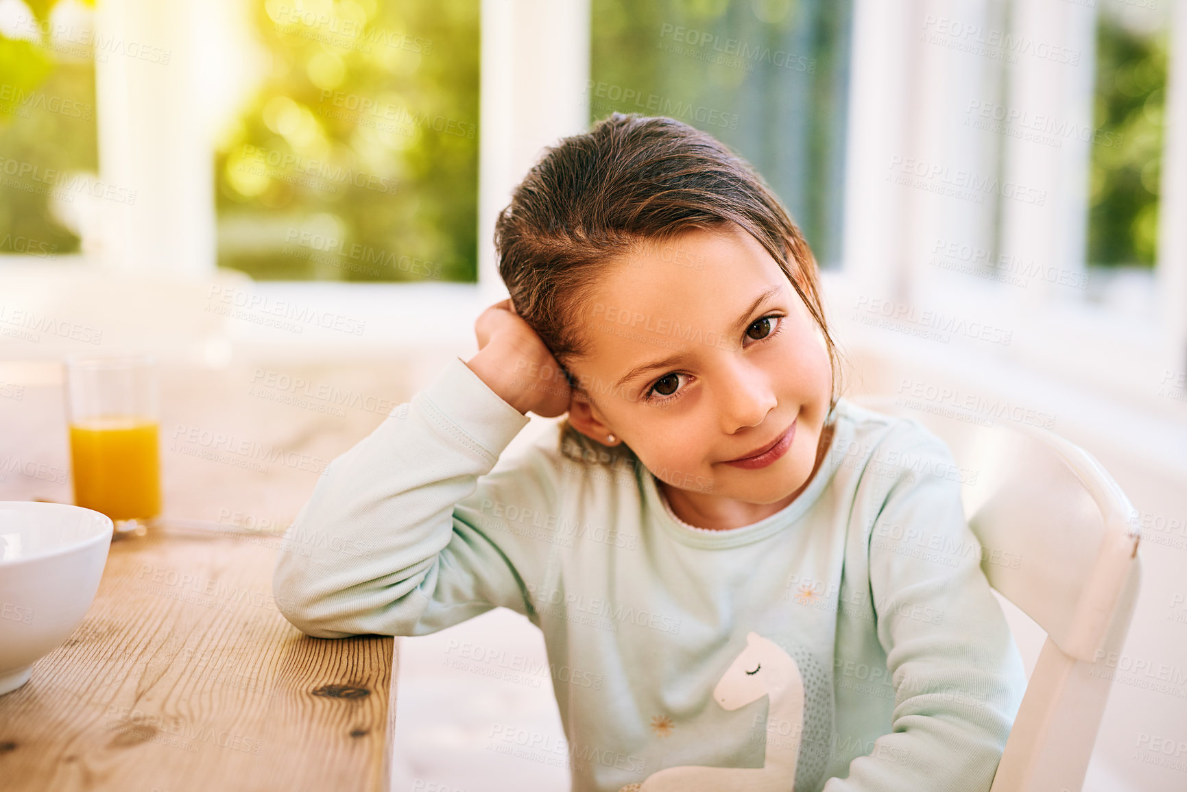 Buy stock photo Portrait of a cheerful little girl sitting next to a kitchen table with her one hand next to her head while looking at the camera