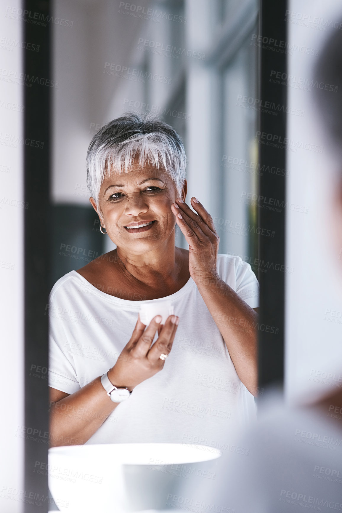 Buy stock photo Shot of a cheerful mature woman applying skin cream on her face while looking into a mirror at home