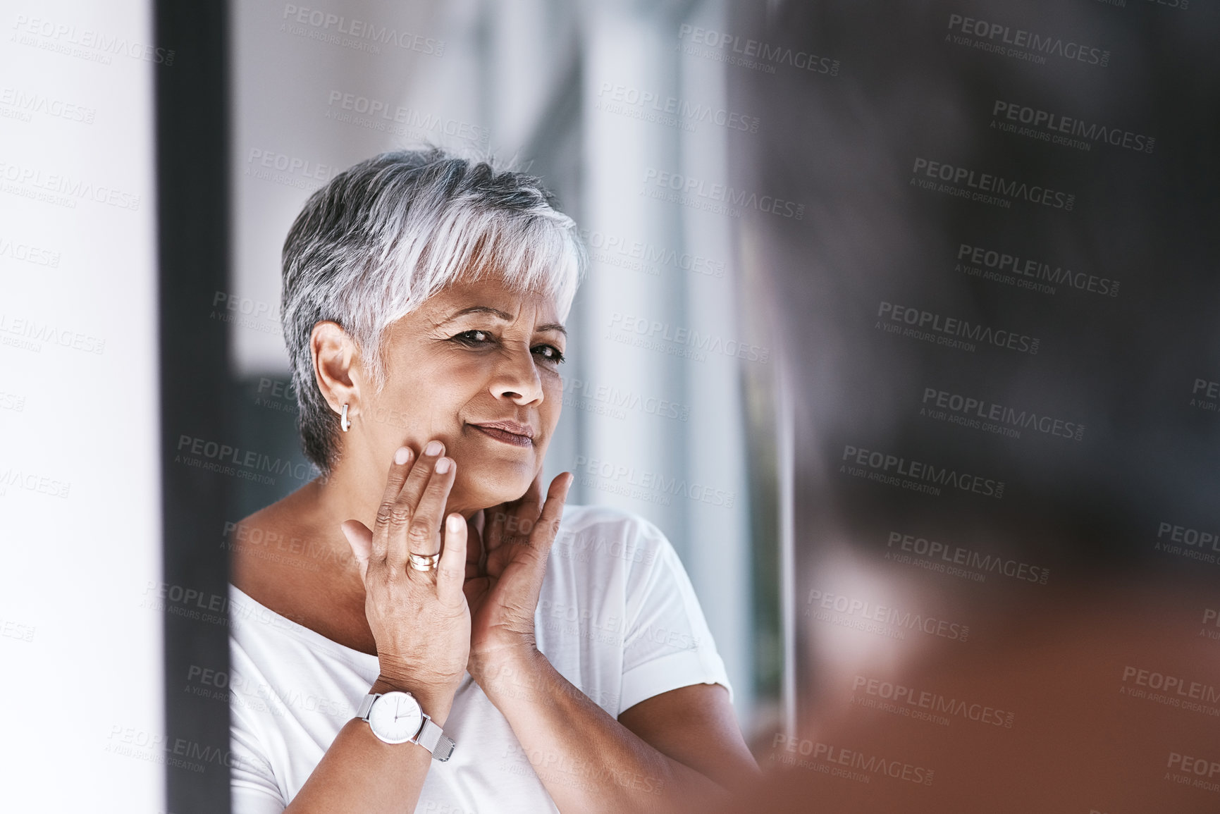Buy stock photo Shot of a cheerful mature woman touching her face with her hands while looking at her reflection in a mirror