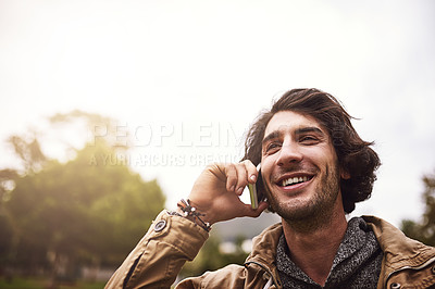 Buy stock photo Shot of a cheerful young man talking on his phone while standing outside in a park