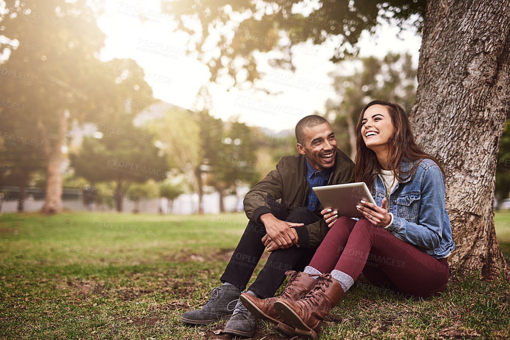 Buy stock photo Shot of a cheerful young  couple sitting down under a tree with a digital tablet outside in a park