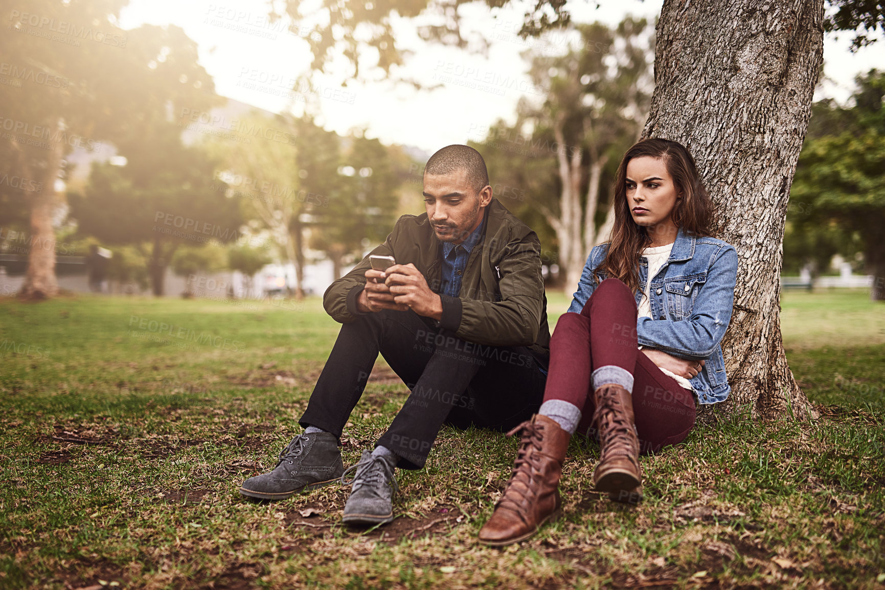 Buy stock photo Shot of a cheerful young man sitting down under a tree with his unhappy girlfriend while using his phone outside in a park