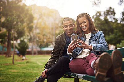 Buy stock photo Shot of a cheerful young couple sitting down on a bench while using a phone together outside in a park