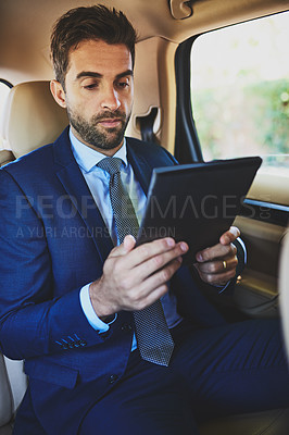 Buy stock photo Shot of a focused young businessman working on his digital tablet while being seated in the back of a car
