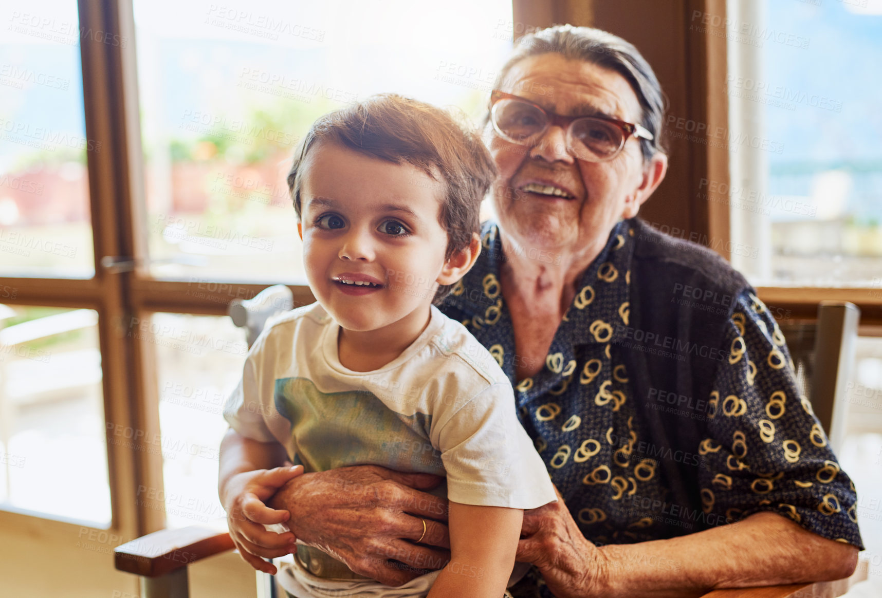 Buy stock photo Portrait of a cheerful little boy sitting on his great grandmother's lap while looking into the camera at home