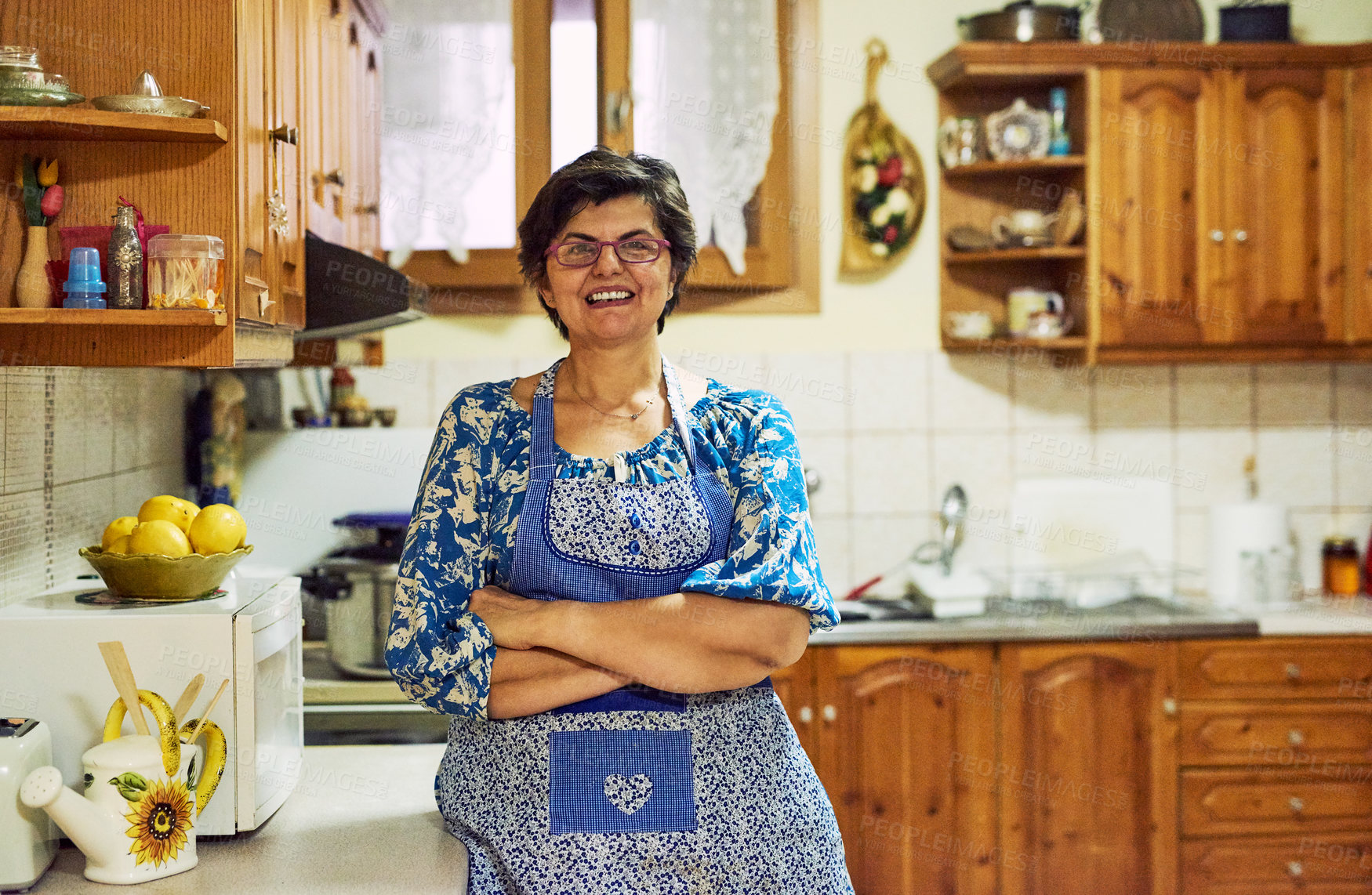 Buy stock photo Portrait of a cheerful mature woman standing with arms folded while looking into the camera in the kitchen at home